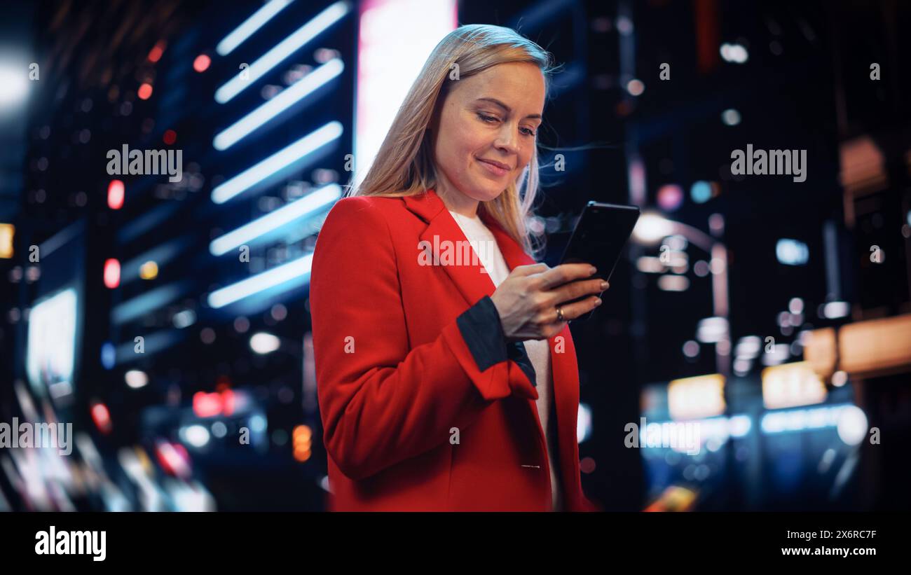 Belle femme debout, en utilisant un smartphone sur la rue de la ville avec Neon Bokeh lumières brillant la nuit. Confiant souriant belle femme à l'aide du téléphone portable. Portrait cinématographique moyen angle bas. Banque D'Images
