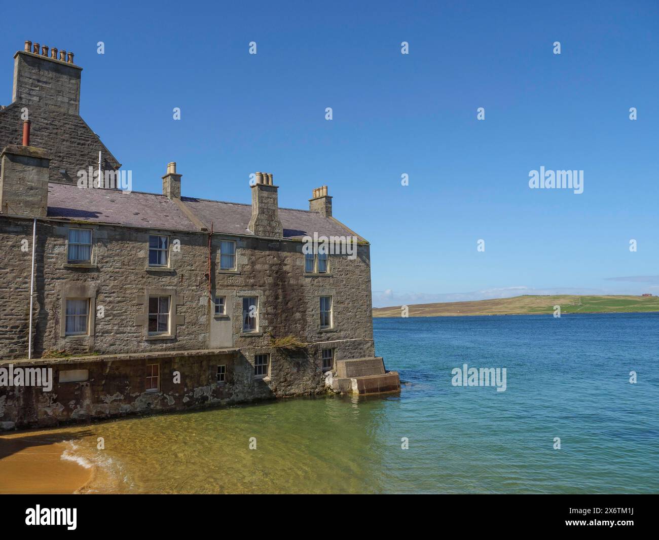 Ancien bâtiment en pierre sur le front de mer avec un ciel clair et une mer calme en arrière-plan, mer calme avec des rochers et des champs avec des maisons historiques, Lerwick Banque D'Images