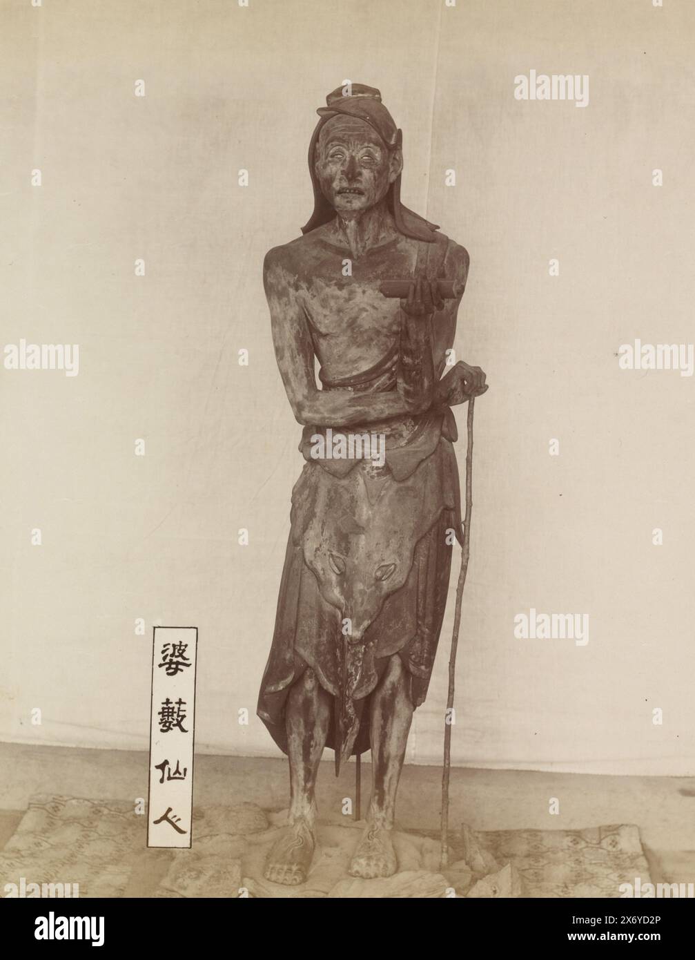 Sculpture des Basuses immortels au temple Sanjusangendo à Kyoto : une figure debout d'un homme mince avec de longs cheveux, tenant un bâton et un rouleau dans sa main gauche, avec un panneau sur le sol à côté de lui qui lit 婆藪仙人, 'Basu sennin' ('le Basu immortel'). Basu (sanskr. Vasu) est représenté comme un ascète indien ; il apparaît dans les temples bouddhistes japonais comme l'un des assistants de Senju Kannon, le Bodhisattva à mille bras Avalokiteshvara., partie de l'album avec 69 photos d'un voyage à travers le Japon., photographie, anonyme, Japon, 1890 - 1894, carton, impression albumine, hauteur, 270 mm × largeur, 204 mm Banque D'Images