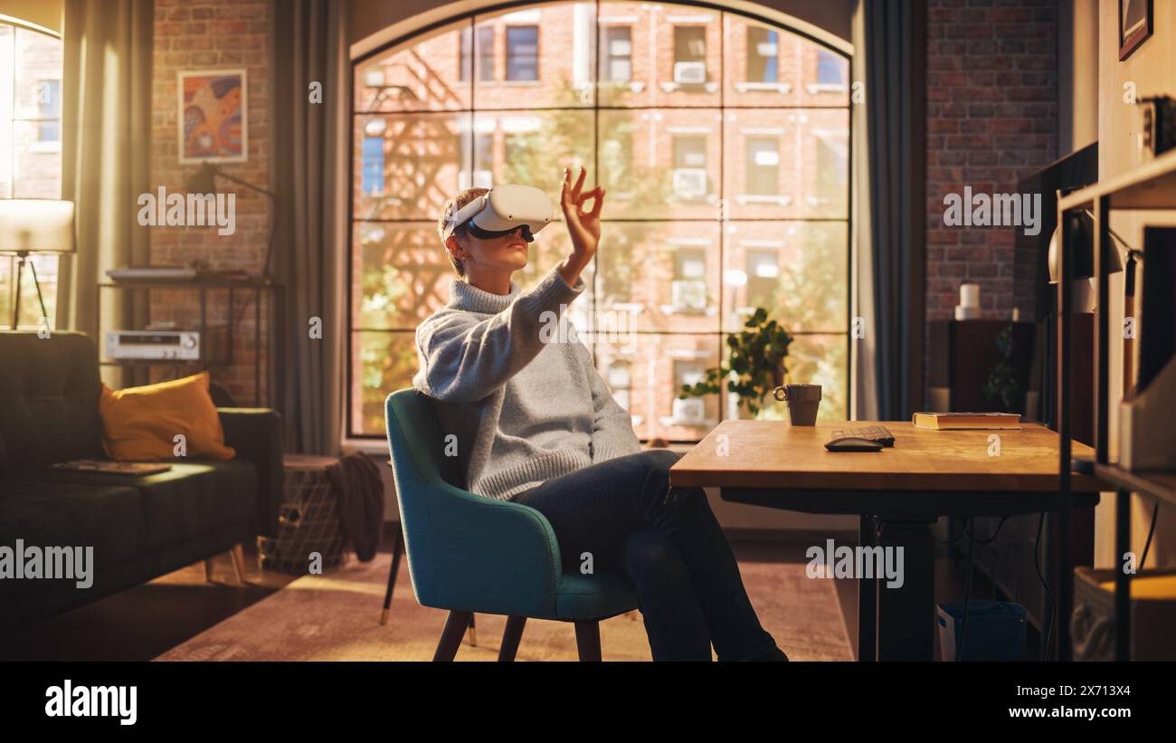 Jeune joueuse féminine avec les cheveux courts s'amusant et jouant au jeu de réalité virtuelle à la maison dans son salon. Elle porte un casque moderne et utilise des contrôleurs. Appartement élégant avec grande fenêtre. Banque D'Images