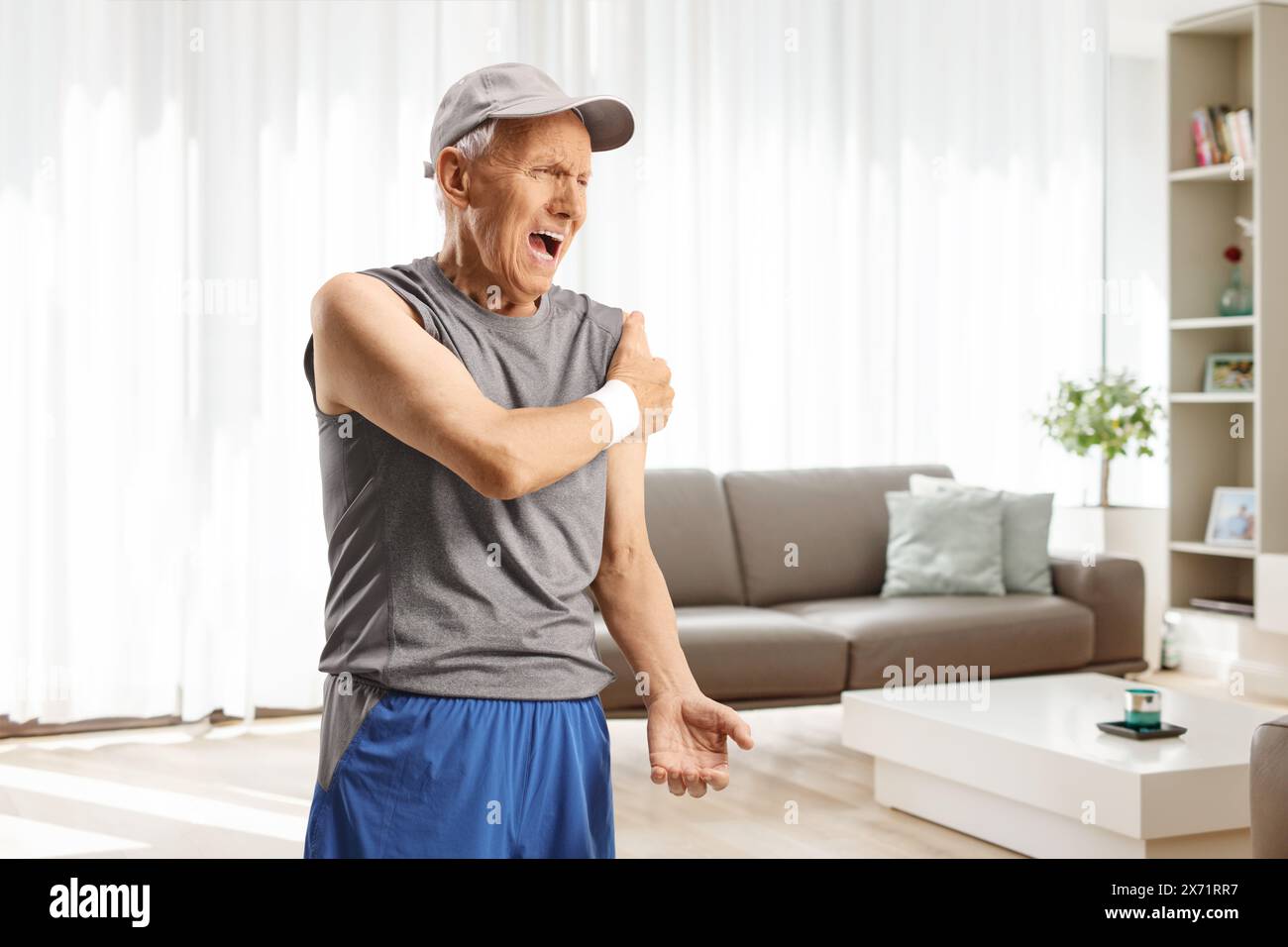 Homme âgé en vêtements de sport debout dans un salon et tenant son épaule, la douleur et le concept de soins de santé Banque D'Images
