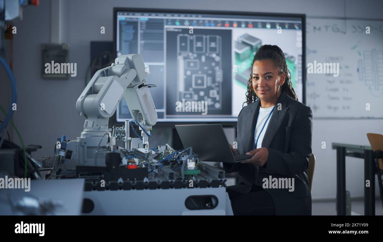 Portrait de Smiling Black Developer à l'aide d'un ordinateur portable tout en travaillant avec un bras robotique dans une salle de classe universitaire moderne. Femme regardant caméra. Concept de réussite et de programmation. Prise de vue moyenne Banque D'Images