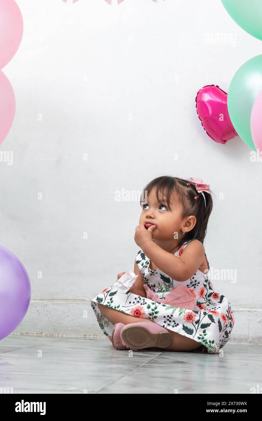 petite brune latina fille assise sur le sol entourée de ballons, mettant une main à sa bouche tout en levant les yeux Banque D'Images