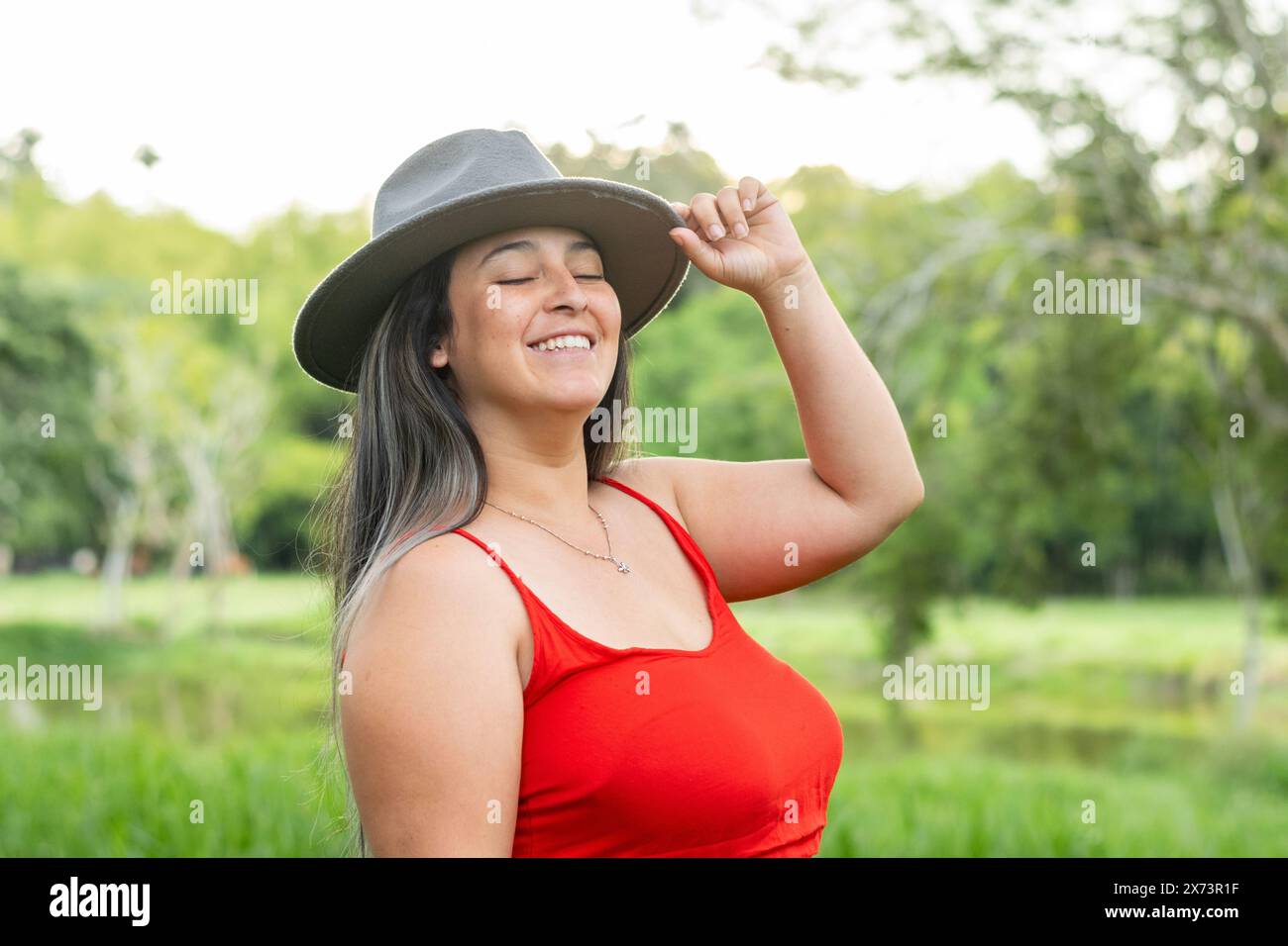 femme latina souriant joyeusement alors qu'elle ferme les yeux et tient son chapeau gris dans sa main Banque D'Images