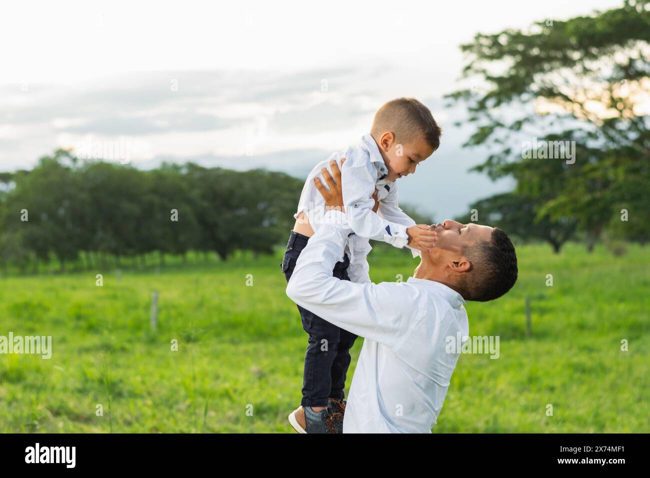 Jeune père latino tenant son jeune fils tout en jouant avec lui dans un champ d'herbe verte. Banque D'Images