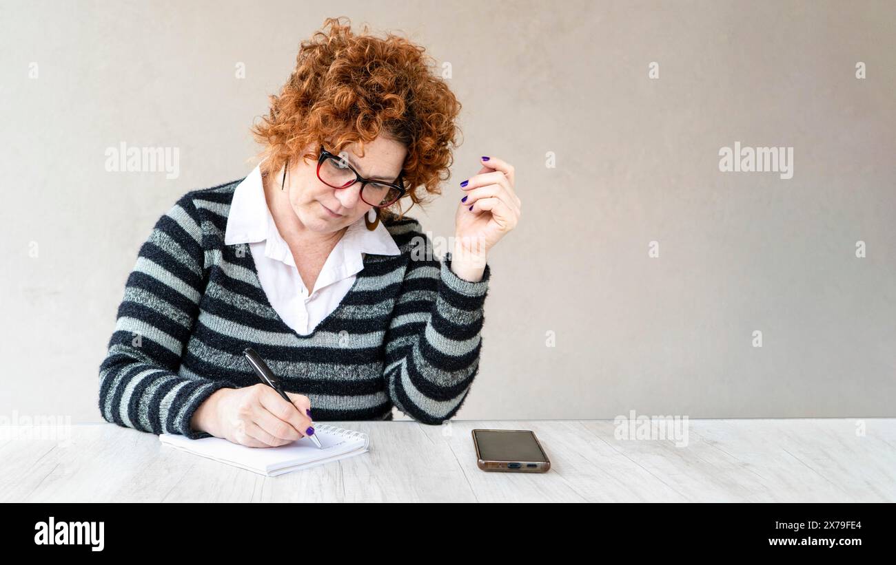Une femme aux cheveux roux et aux lunettes écrit dans un cahier. Elle a un téléphone portable sur la table devant elle Banque D'Images