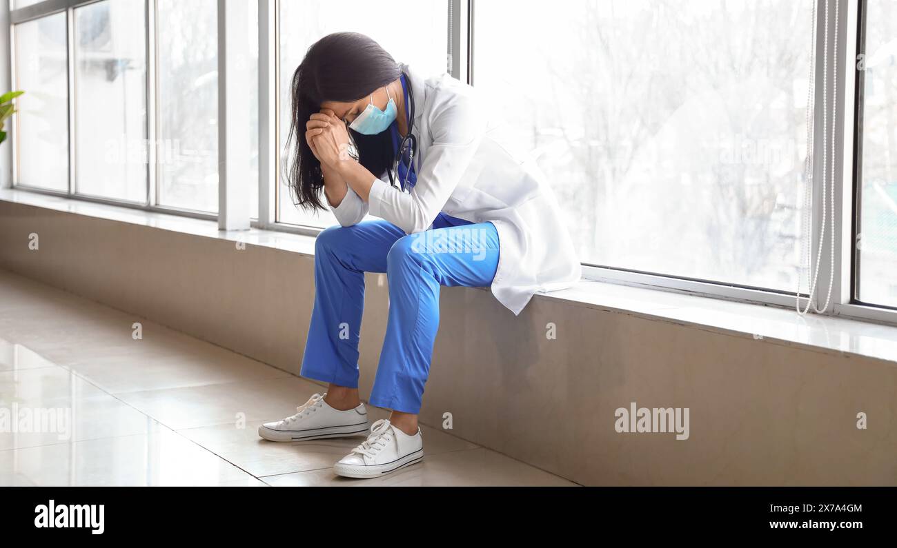 Médecin féminin inquiet assis sur le rebord de la fenêtre dans le couloir de la clinique Banque D'Images