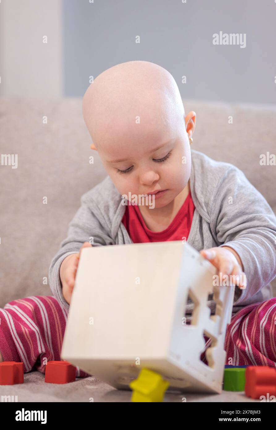 Bébé fille avec le cancer et la perte de cheveux due à la chimiothérapie jouant avec le jouet en bois sur le canapé Banque D'Images