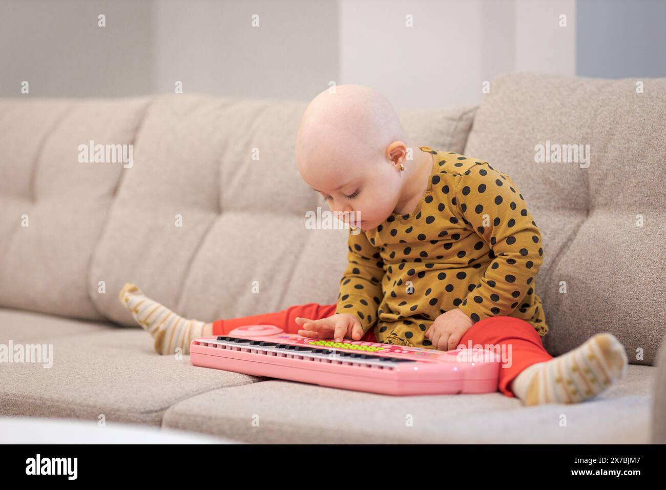 Bébé fille avec cancer et perte de cheveux due à la chimiothérapie jouant avec un piano sur le canapé Banque D'Images