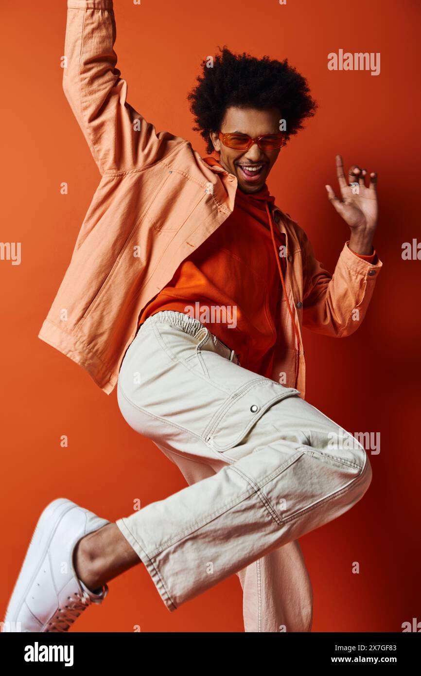 Homme afro-américain bouclé dans une tenue tendance et des lunettes de soleil, sautant énergiquement dans l'air sur fond orange. Banque D'Images