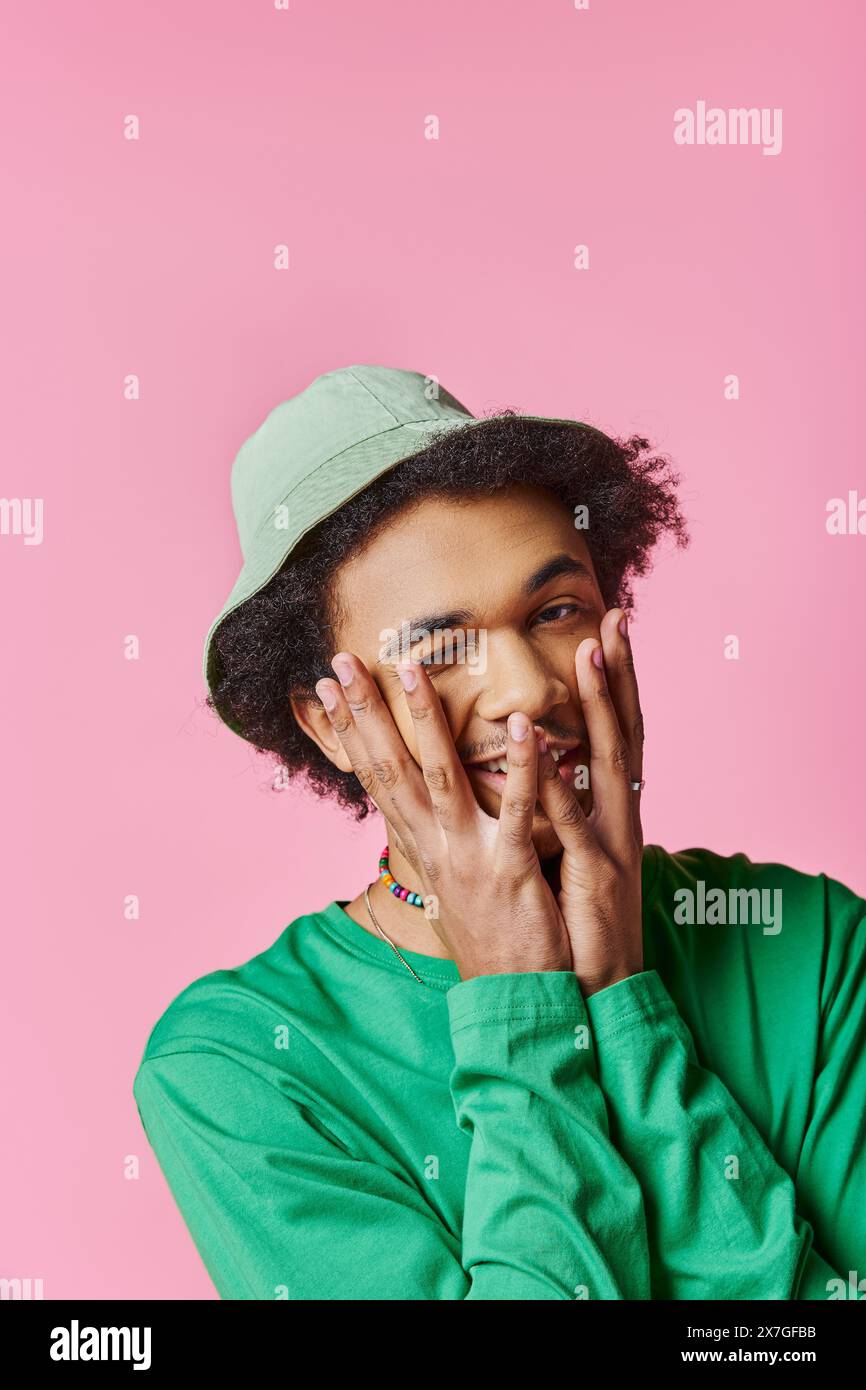 Un jeune homme afro-américain frisé et joyeux porte une chemise verte et un chapeau sur un fond rose. Banque D'Images