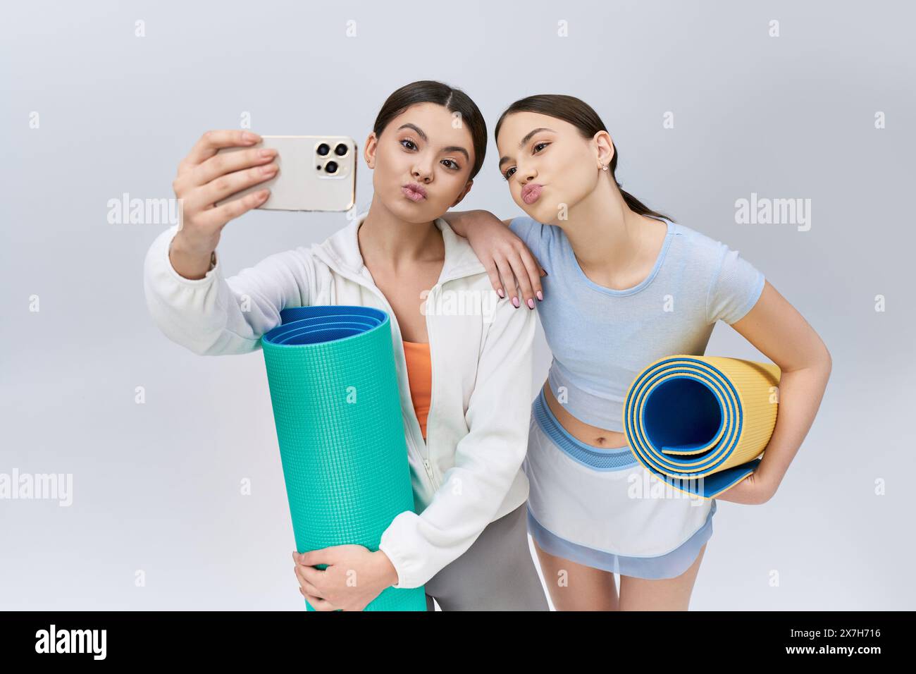 Deux jolis amis adolescents, aux cheveux bruns et aux vêtements de sport, capturent un selfie avec un tapis de yoga dans un cadre de studio. Banque D'Images