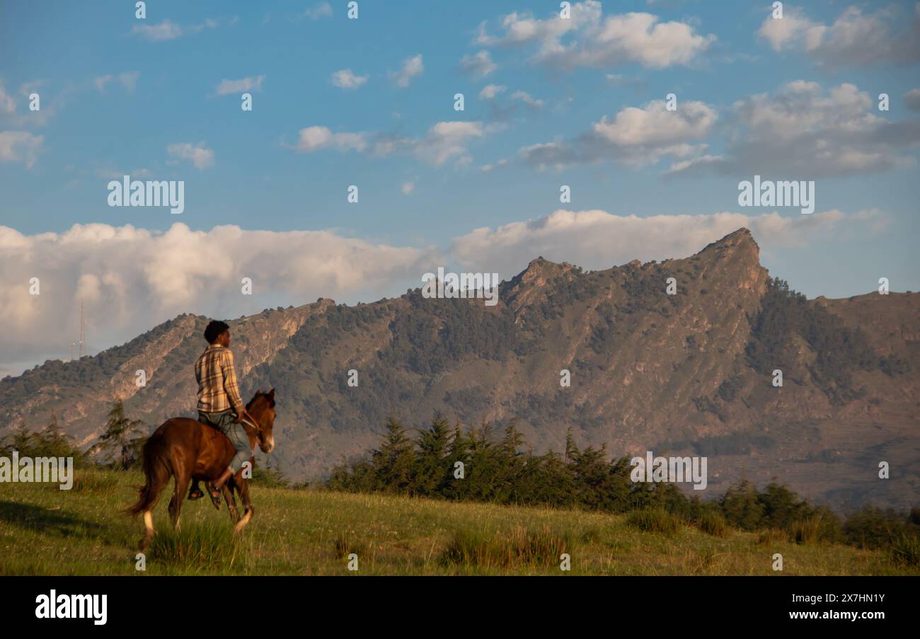 Un Africain monte à cheval dans une vallée sereine et verdoyante, entourée de paysages à couper le souffle. Les montagnes majestueuses dominent l'arrière-plan Banque D'Images