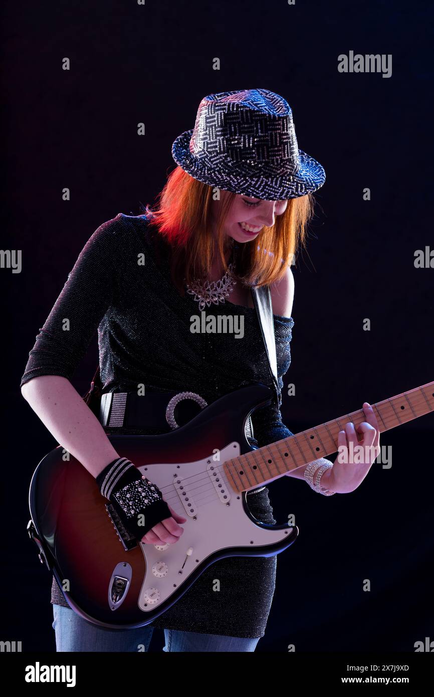 Jeune femme souriante avec des cheveux auburn joue de la guitare électrique sur scène. Elle porte un chapeau étincelant, un haut à paillettes foncées, un Jean et des gants sans doigts, p. ex. Banque D'Images