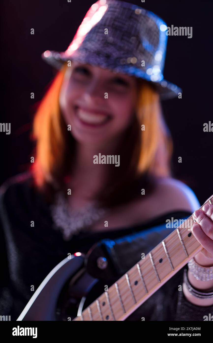 Une jeune femme aux cheveux auburn, dans un chapeau étincelant et un haut à paillettes foncées, joue de la guitare électrique. Son sourire et sa pose confiante sur scène transmettent son orl Banque D'Images