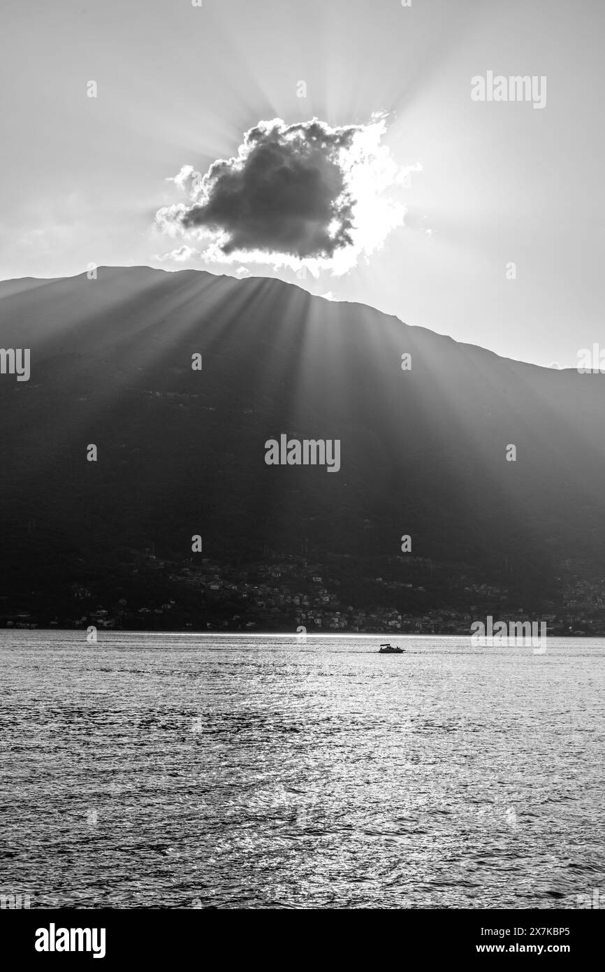 Une scène tranquille capturant la beauté sereine d'un lac de montagne au coucher du soleil. Les rayons du soleil traversent un nuage, projetant des faisceaux lumineux et des reflets étincelants sur la surface de l'eau. Un bateau solitaire profite de l'atmosphère paisible, entouré par la silhouette des montagnes. Image en noir et blanc. Banque D'Images