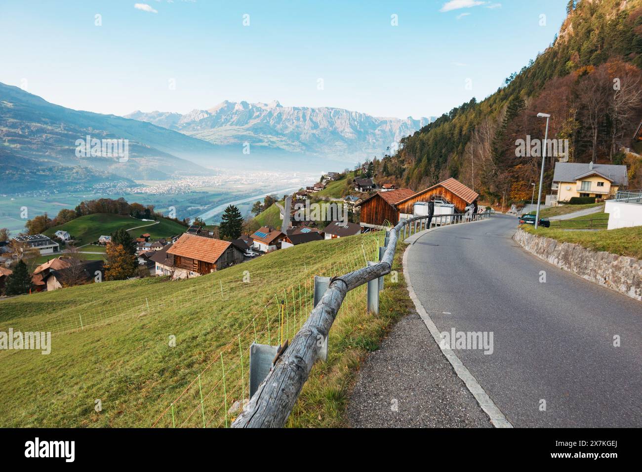 Une route sinueuse mène à travers un village pittoresque du Liechtenstein niché dans les contreforts des Alpes, avec une vue sur une vallée et des montagnes lointaines Banque D'Images