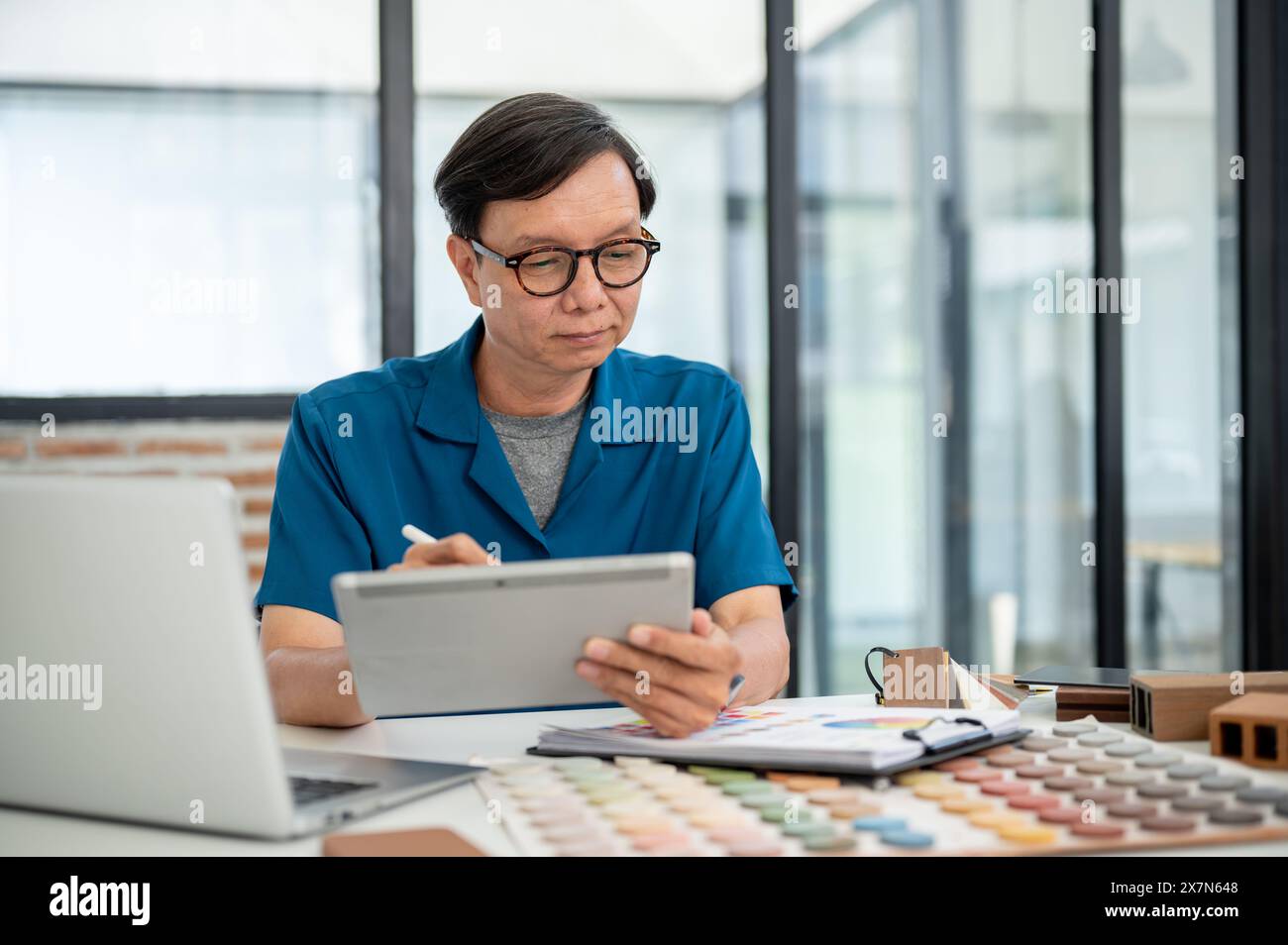 Un designer d'intérieur professionnel masculin asiatique est en train de concevoir l'intérieur du bâtiment sur sa tablette numérique, travaillant à son bureau dans son studio de bureau. Banque D'Images