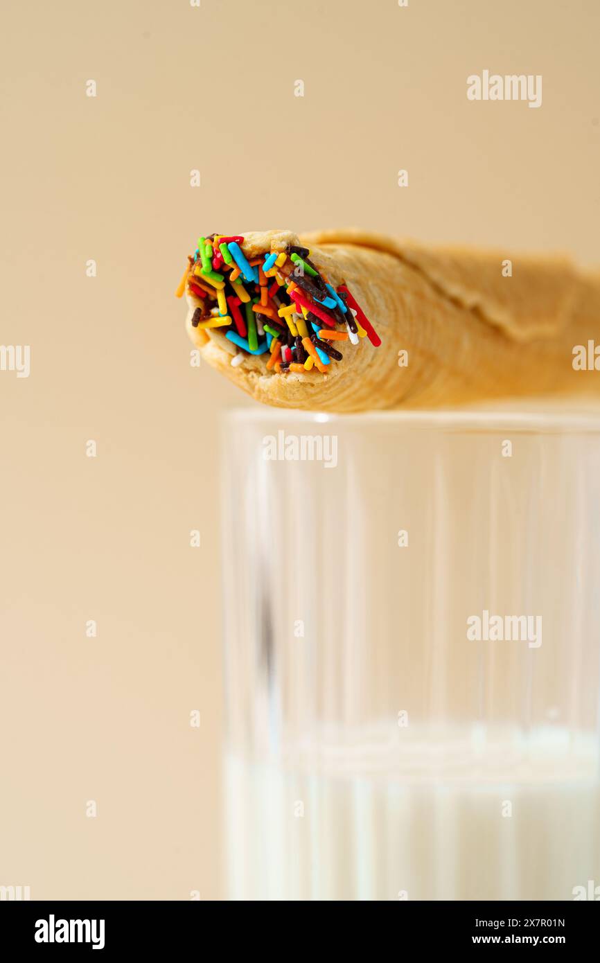 Image en gros plan d'un rouleau de gaufres surmonté de saupoudrages colorés reposant sur le bord d'un verre de lait sur un fond beige. Banque D'Images