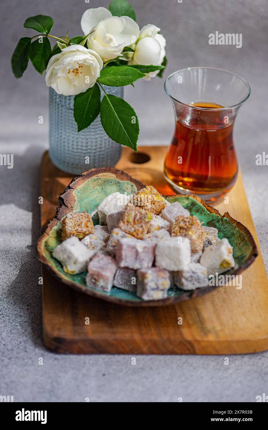 Un assortiment de délices turcs traditionnels colorés sur une assiette avec un verre de thé noir, complété par un vase de roses blanches fraîches Banque D'Images