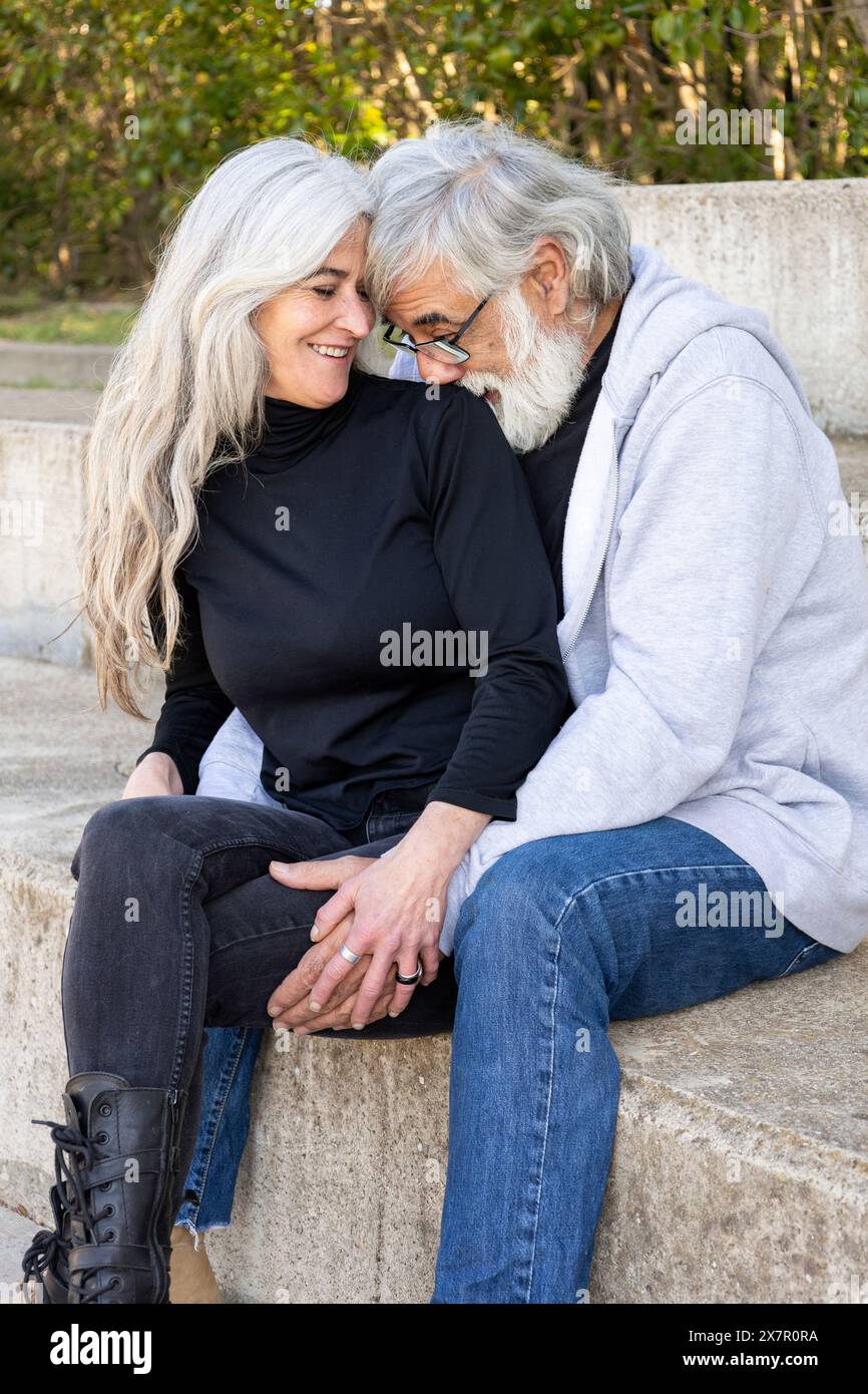 Un couple de personnes âgées affectueux assis étroitement ensemble sur les marches extérieures, exprimant amour et compagnie avec des sourires doux et des mains entrelacées Banque D'Images