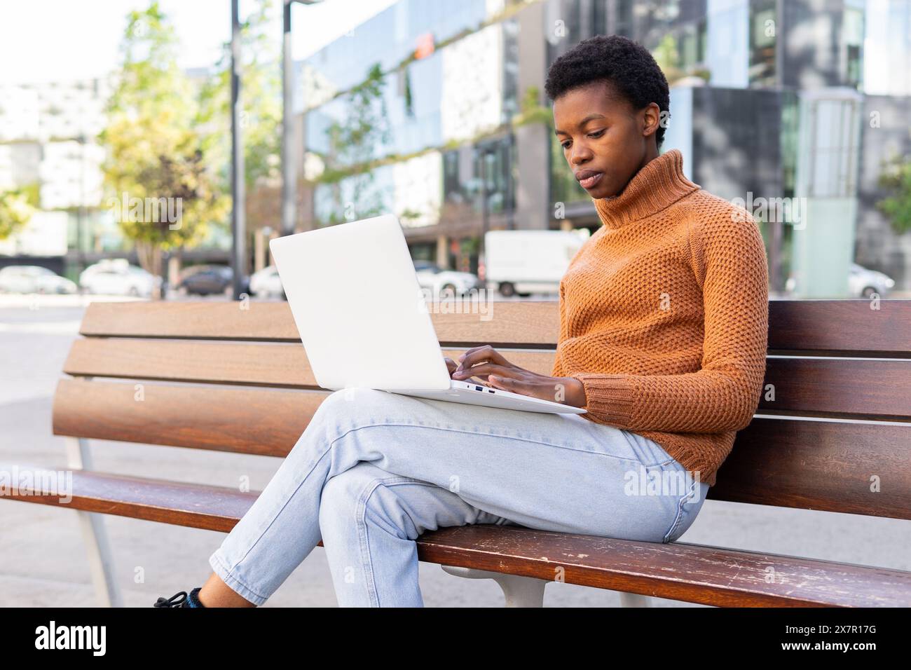 Une étudiante afro-américaine concentrée travaille sur son ordinateur portable blanc tout en étant assise sur un banc à l'extérieur, profondément absorbée par sa tâche. Banque D'Images