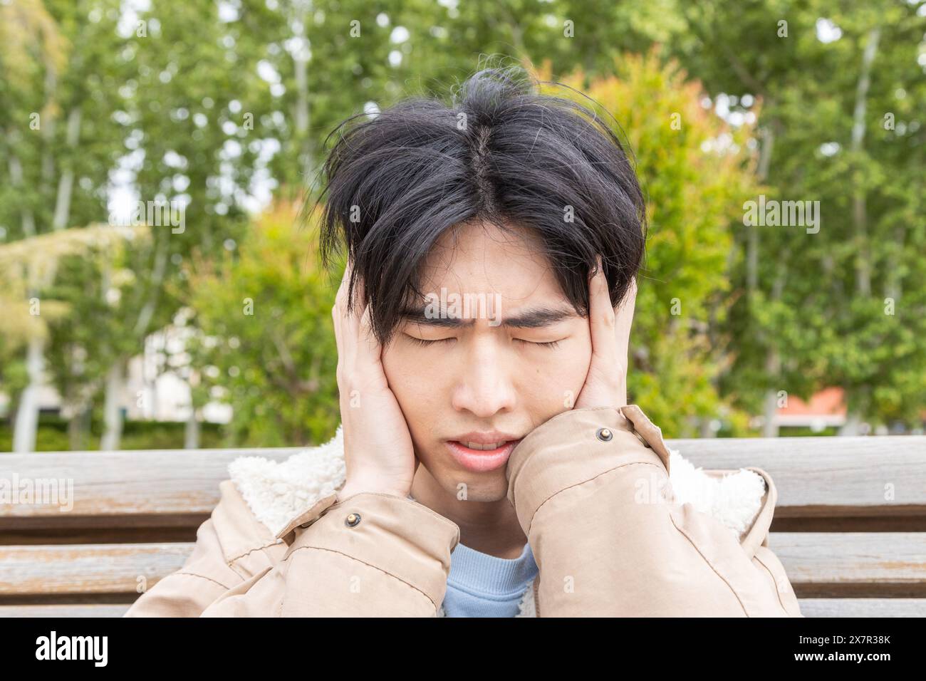 Un jeune homme asiatique est assis sur un banc de parc serrant sa tête dans l'inconfort, dépeignant une scène liée à des symptômes d'hypoglycémie Banque D'Images