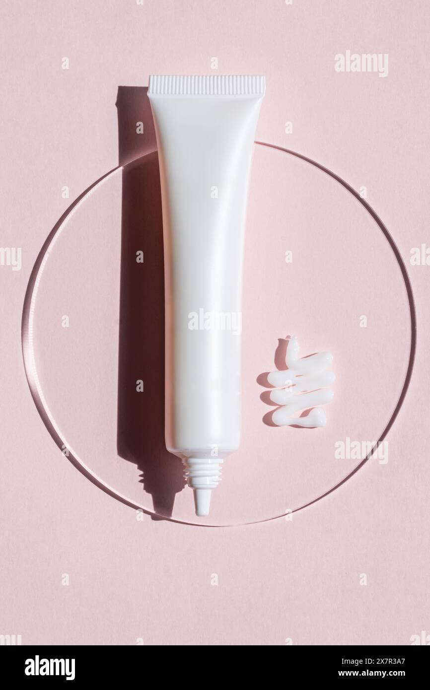 Un tube de crème cosmétique placé à côté de sa cuillère de produit enduit artistiquement sur une surface rose, réfléchissant sur un miroir circulaire. Banque D'Images