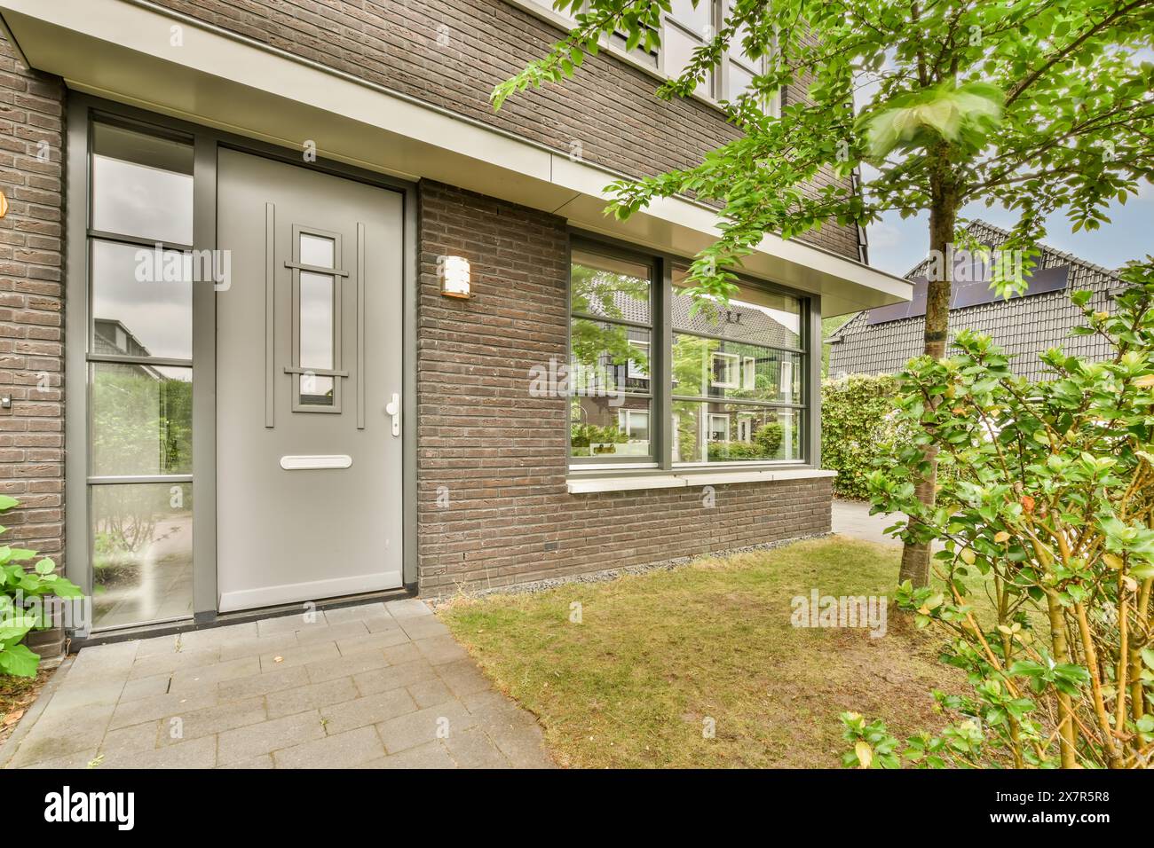 La façade d'une maison de banlieue contemporaine présente une élégante porte d'entrée grise, de grandes fenêtres et des murs de briques rouges, entourés de verdure Banque D'Images