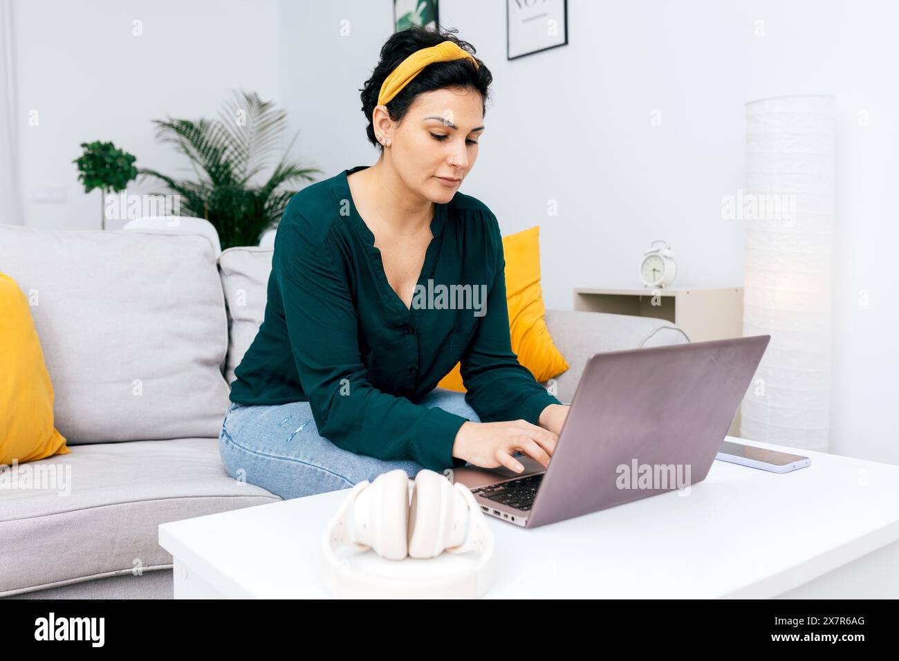 Une femme concentrée dans une blouse vert foncé s'engage avec son ordinateur portable tout en s'asseyant confortablement sur un canapé blanc dans un salon lumineux, orné de M. Banque D'Images