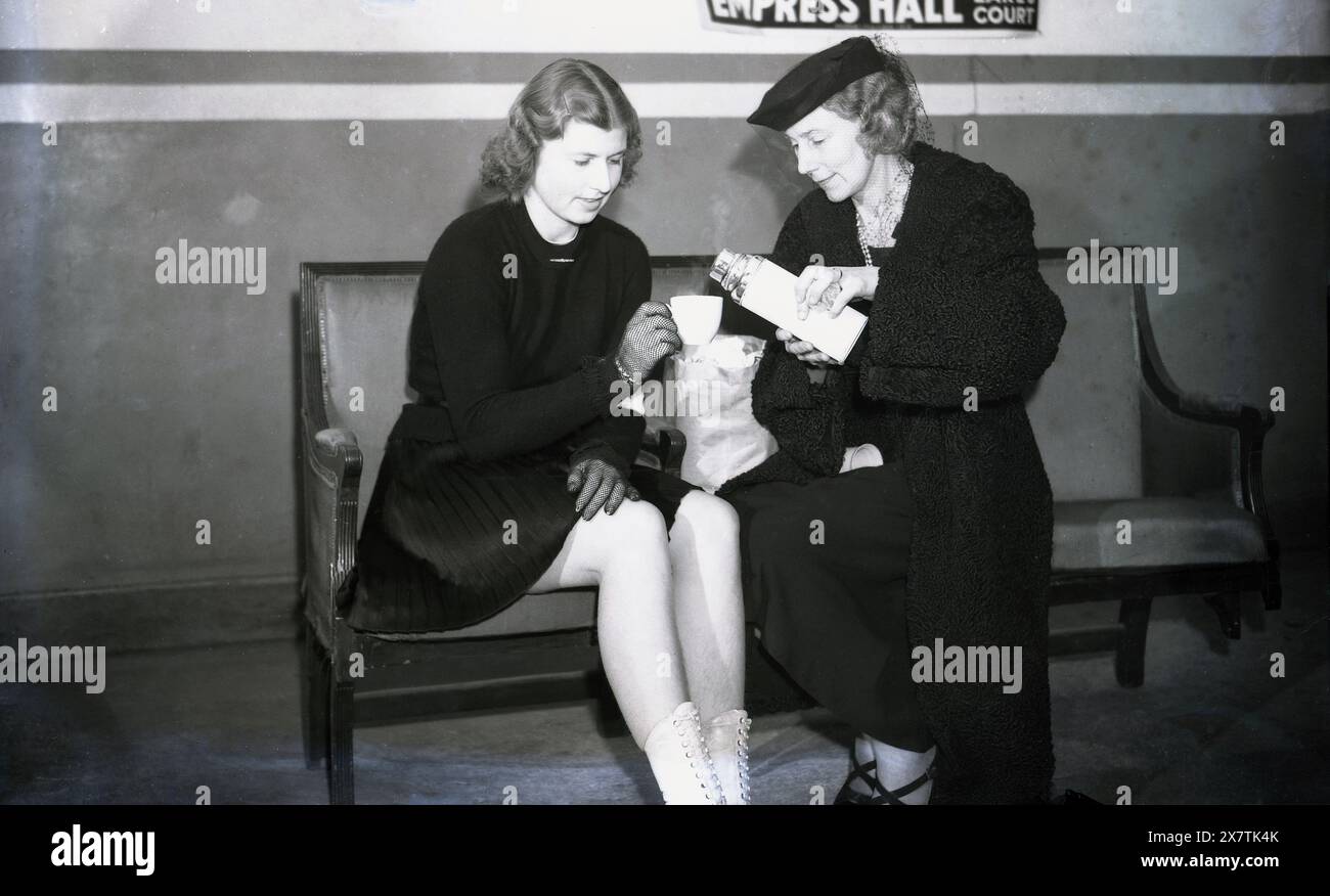 1939, historique, assise sur un banc, la championne européenne en titre de patinage artistique Cecilia Colledge reçoit une boisson chaude par sa mère dans une bouteille à l'Empress Hall, Earls court, Londres, Angleterre, Royaume-Uni. La salle était le lieu de l'épreuve féminine des Championnats d'Europe de patinage artistique 1939, remportés par Colledge sur une autre patineuse britannique, Megan Taylor. Banque D'Images