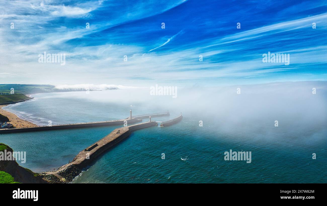 Vue aérienne d'une jetée côtière s'étendant dans l'océan avec un phare à l'extrémité. Le ciel est clair avec quelques nuages, et il y a un brouillard qui roule Banque D'Images