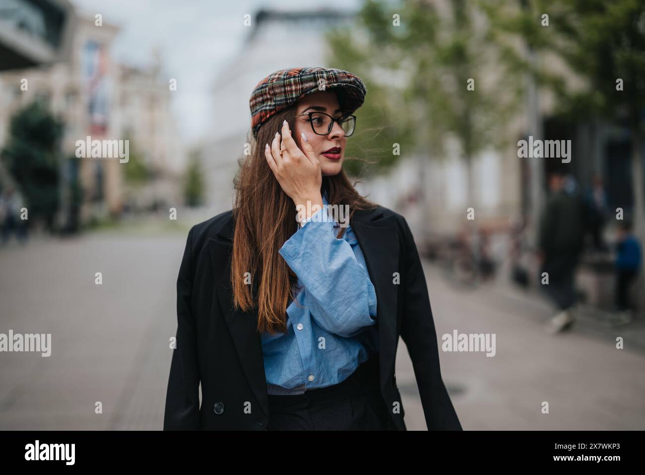 Femme confiante dans une tenue élégante marchant dans un cadre urbain portant un chapeau et des lunettes Banque D'Images