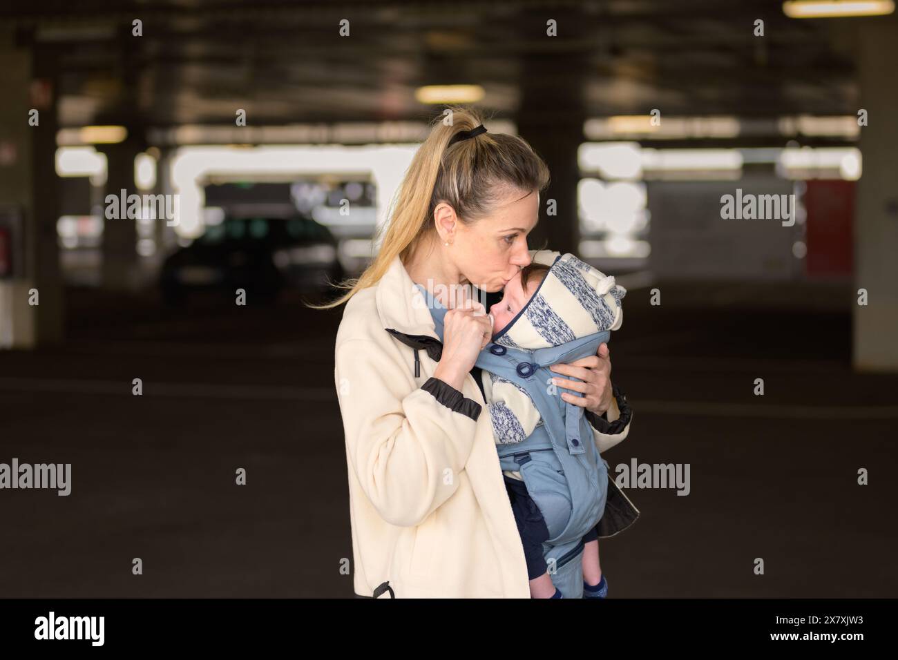 Vue latérale d'une femme heureuse embrassant son bébé tout en le tenant et le portant dans un porte-bébé dans un parking Banque D'Images