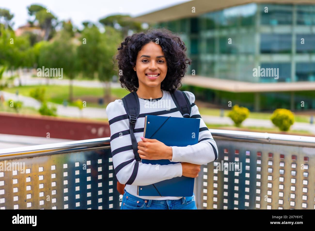 Portrait d'une étudiante latine souriante avec des vêtements décontractés et sac à dos debout fièrement avec dossier à l'extérieur de l'université Banque D'Images