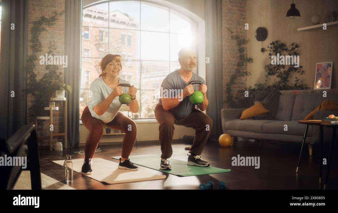 Heureux couple Senior faisant des exercices du matin et Kettlebell entraînement ensemble à la maison dans le salon ensoleillé. Mode de vie sain, remise en forme, loisirs, objectifs de couple, bien-être et retraite. Banque D'Images