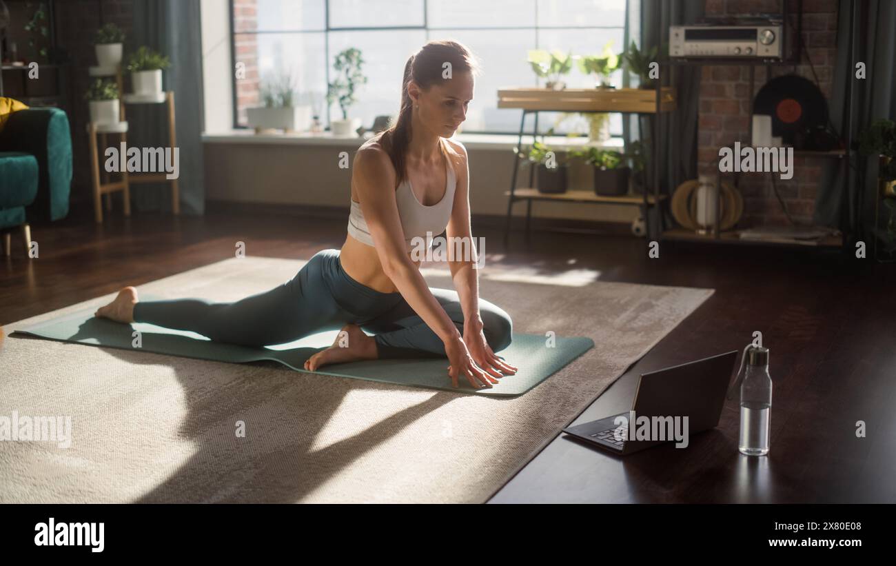 Athlétique jeune femme exerçant, étirant et faisant du yoga tout en regardant la vidéo d'entraînement sur un ordinateur portable. Belle femme en vêtements de sport pratiquant différentes poses Asana sur le tapis. Banque D'Images