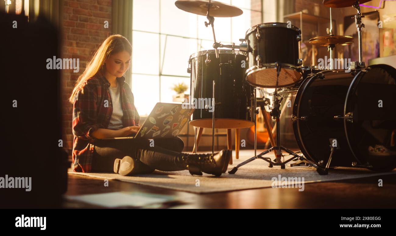 Jeune femme à l'aide d'un ordinateur portable, travaillant à partir d'un salon à domicile avec un ensemble de batterie. Fille alternative assise sur le sol, vérifiant les médias sociaux et faisant du travail à distance. Artiste musical à succès à la maison. Banque D'Images