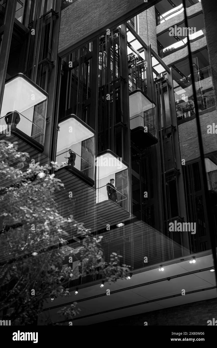 Image monochrome de trois ascenseurs en verre illuminés chacun avec une personne d'affaires à l'intérieur et à l'intérieur d'un immeuble de bureaux moderne Banque D'Images