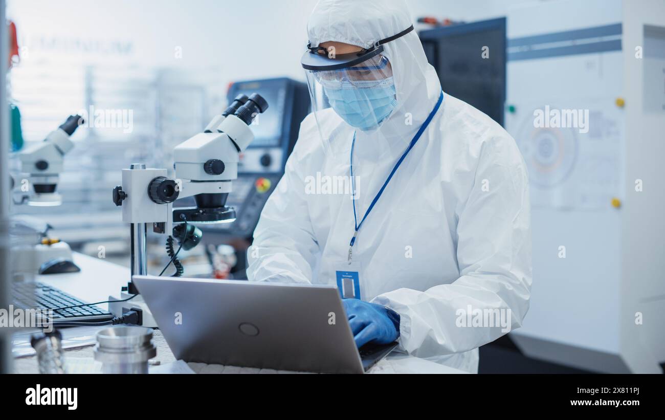 Usine de fabrication électronique : ingénieur en combinaison stérile travaillant sur un ordinateur portable, examinant une carte de circuit imprimé avec des micropuces à travers un microscope et testant de nouveaux équipements électriques. Banque D'Images