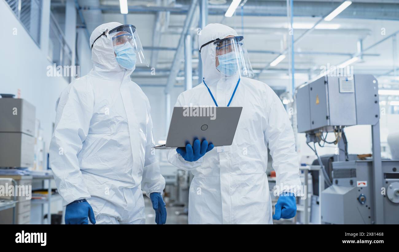 Deux scientifiques marchant dans une usine industrielle lourde dans des combinaisons stériles et des masques faciaux, en utilisant un ordinateur portable. Examen des paramètres de la machine industrielle et configuration de la fonctionnalité de production. Banque D'Images