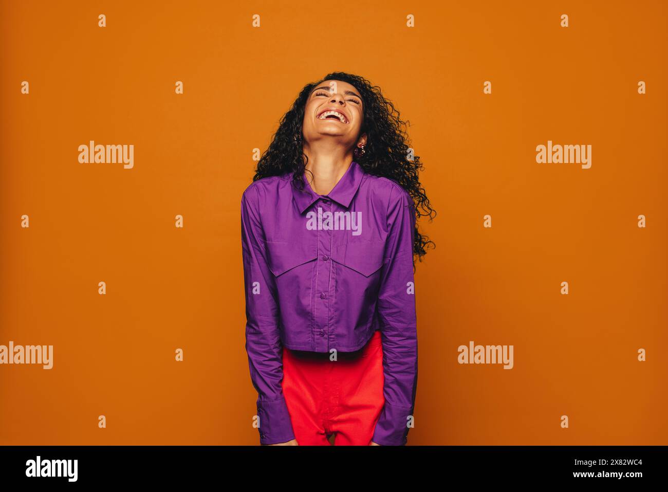 Femme joyeuse avec des cheveux bouclés sur un fond orange vibrant. Portant des vêtements décontractés élégants, elle rayonne d'excitation, de bonheur et de plaisir. Banque D'Images