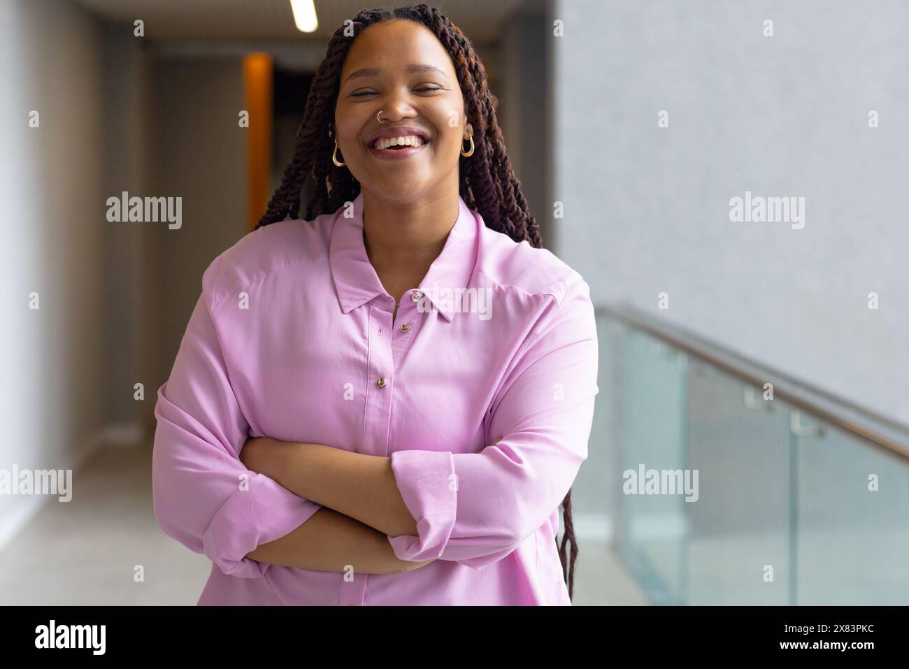 Au bureau moderne pour les affaires, jeune femme biraciale se tient souriante avec les bras croisés Banque D'Images
