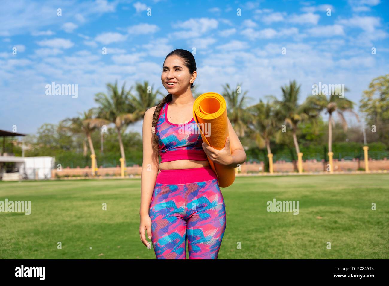 Fit jeune femme indienne tenant tapis de yoga dans le jardin souriant. exercice, méditation, entraînement, extérieur, santé mentale, bien-être. Banque D'Images