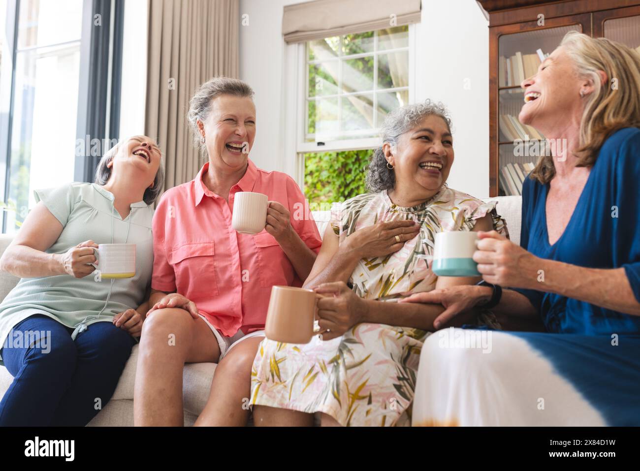 Diverses amies seniors tenant des tasses, riant ensemble à la maison Banque D'Images