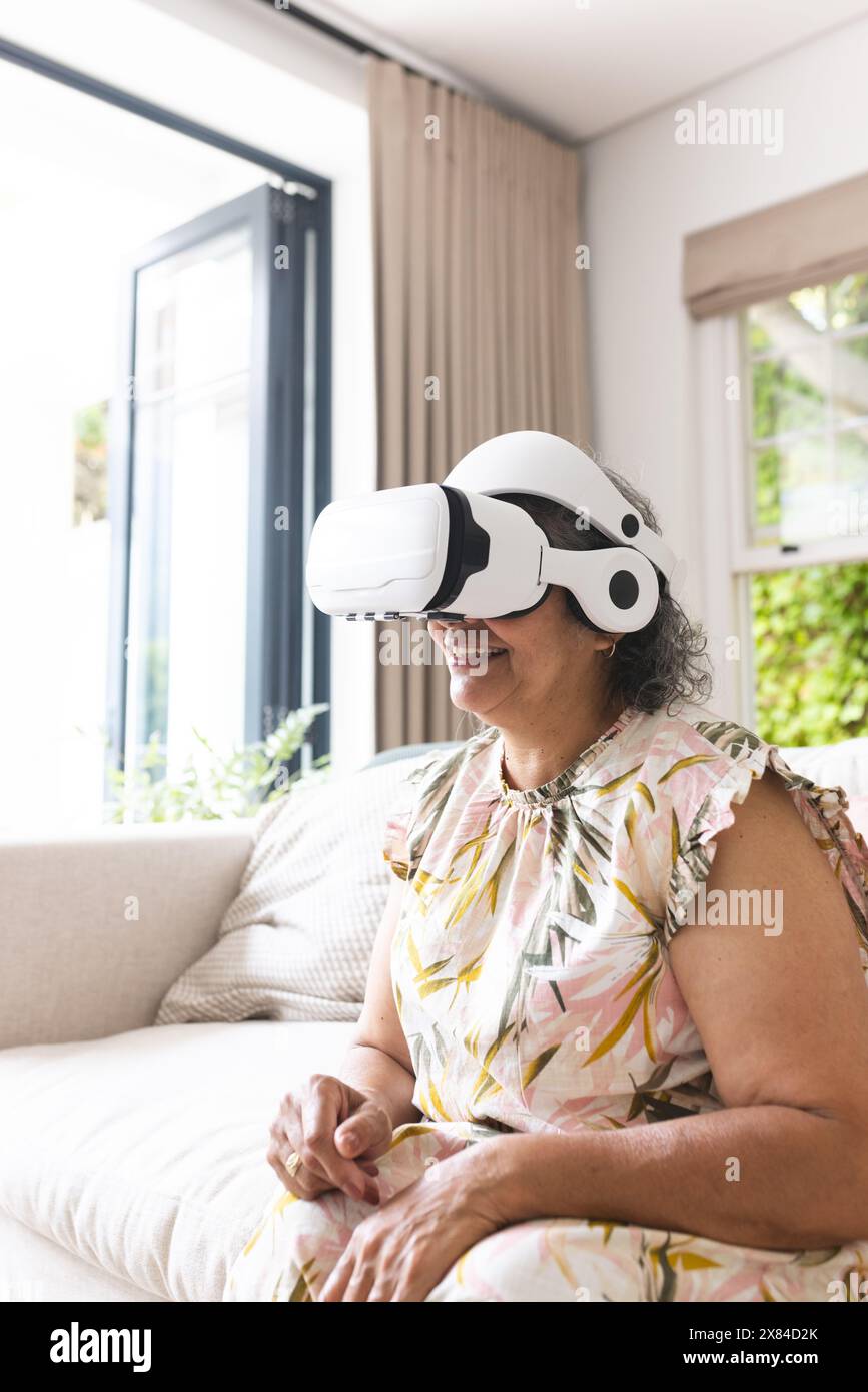 Femme biraciale senior portant un casque de réalité virtuelle à la maison, assise sur le canapé Banque D'Images