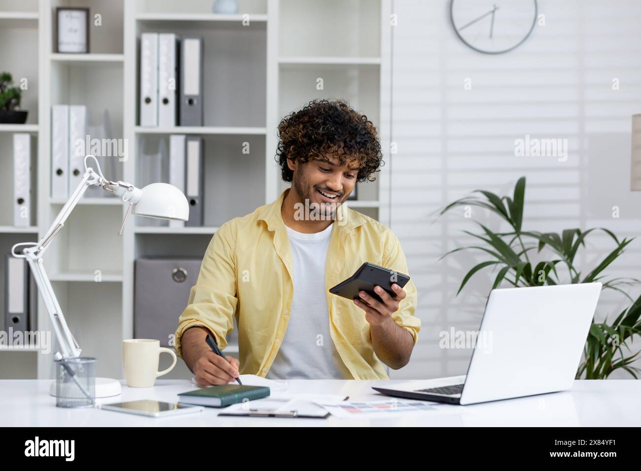 Jeune homme souriant travaillant à la maison avec une tablette et un ordinateur portable, tenant un stylo. Environnement de travail à distance détendu et productif avec fournitures de bureau et mobilier moderne. Banque D'Images