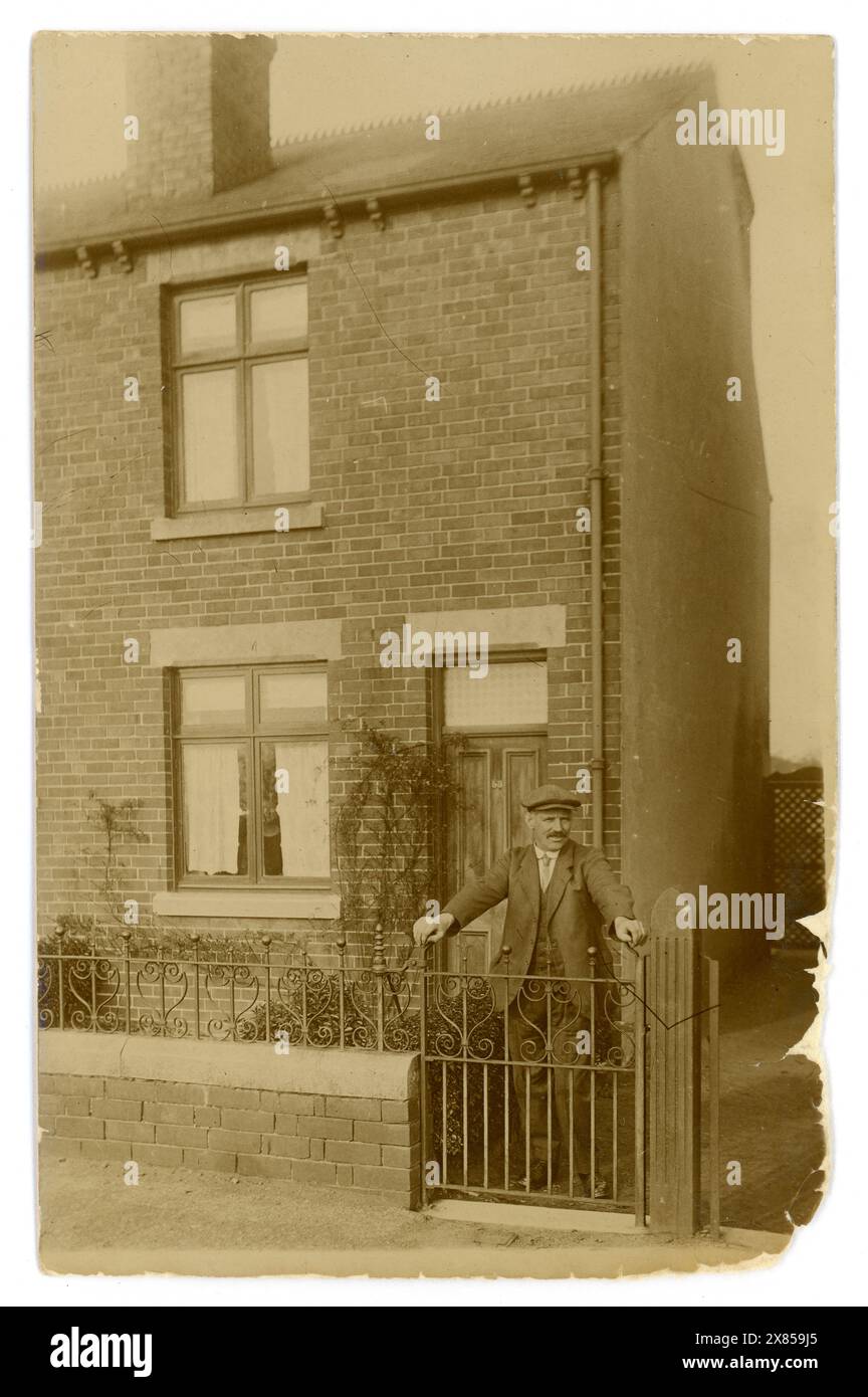 Original WW1 ère carte postale d'homme de la classe ouvrière portant un chapeau plat typique et costume, debout devant la porte d'entrée de sa maison mitoyenne typique en briques rouges britannique no.53, typique logement britannique des années 1900, près de sa porte de jardin, une femme regarde par la fenêtre entre les rideaux, vers 1917, Royaume-Uni Banque D'Images