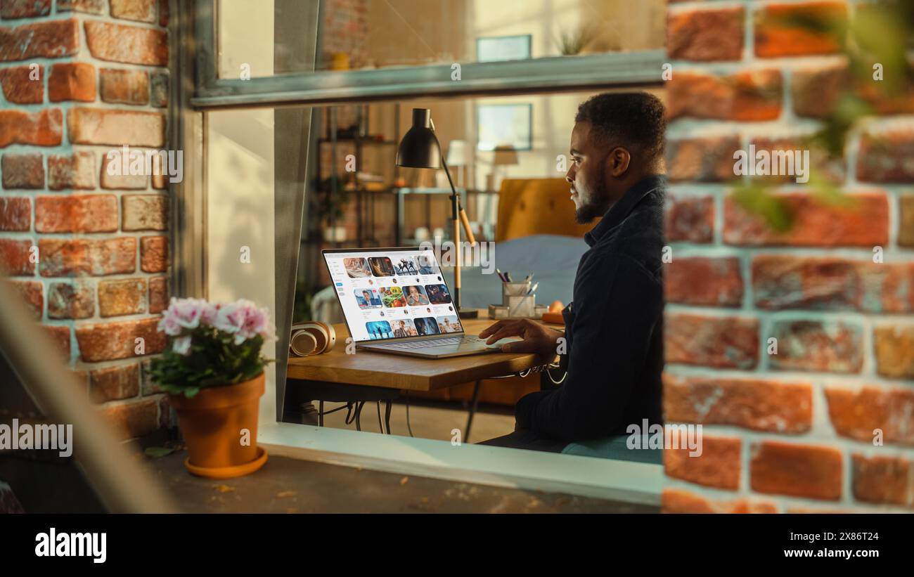 Homme noir utilisant un ordinateur portable avec affichage de plate-forme de streaming vidéo tout en étant assis derrière une table dans un salon élégant. Influenceur naviguant sur Internet. Prise de vue de l'extérieur de la fenêtre. Banque D'Images