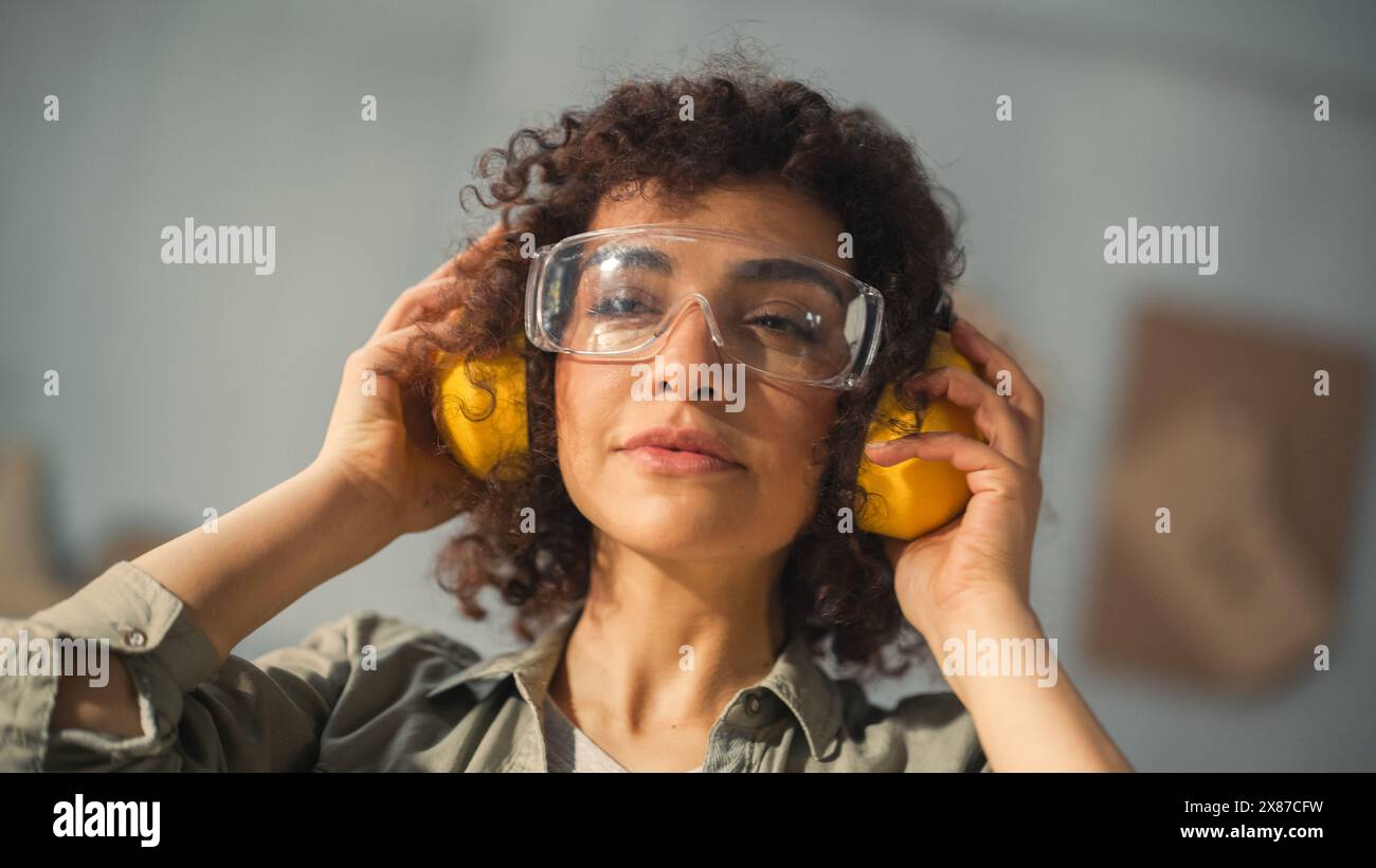 Portrait d'une belle femme multiethnique avec des cheveux bouclés et des yeux bruns mettant sur des lunettes de sécurité protectrices et casque antibruit de construction dans un environnement d'atelier. Banque D'Images